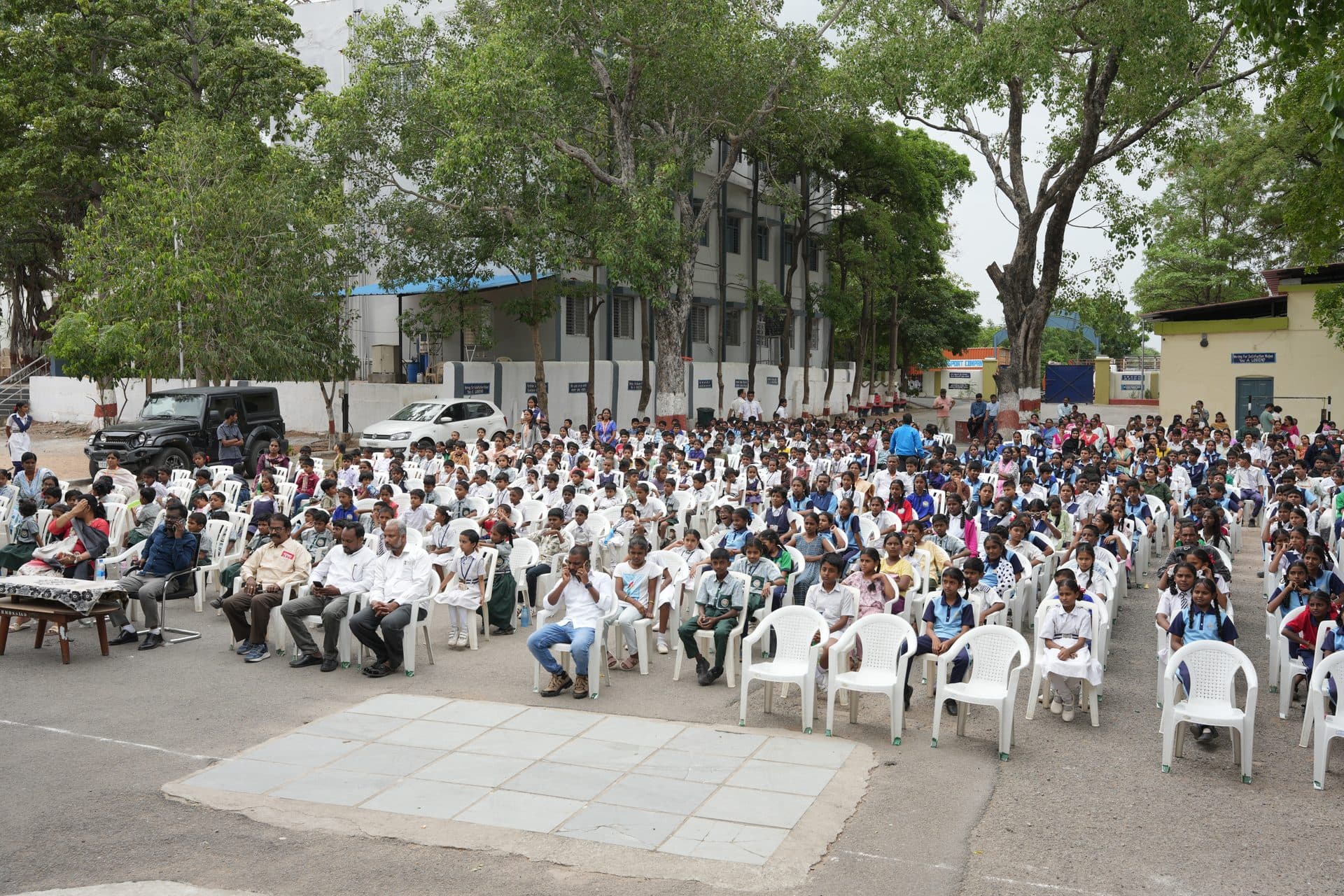Hundreds of students seated in rows of chairs on the school grounds for an open-air programme