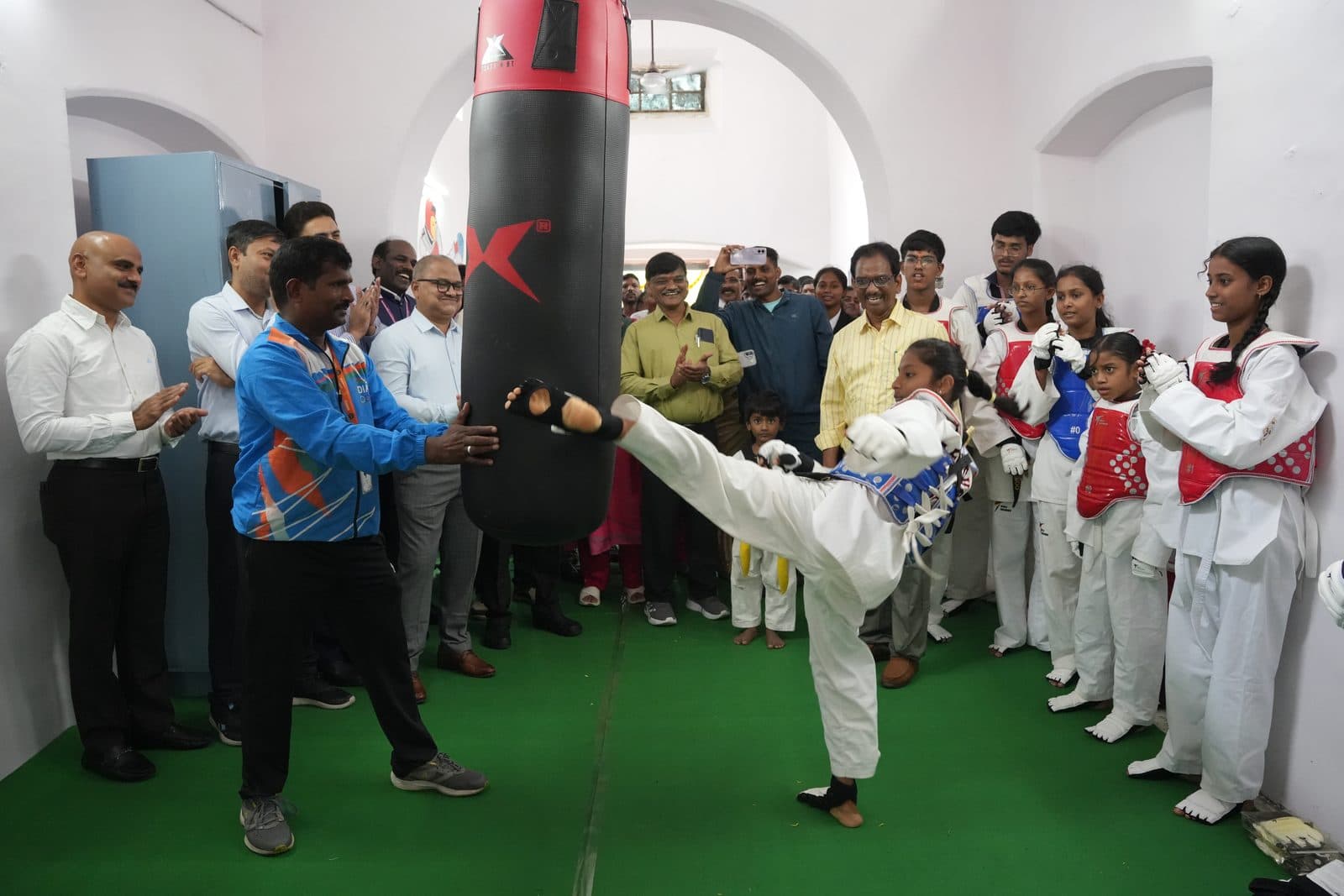 A student in a Taekwondo uniform delivering a high kick to a training bag as classmates and guests watch