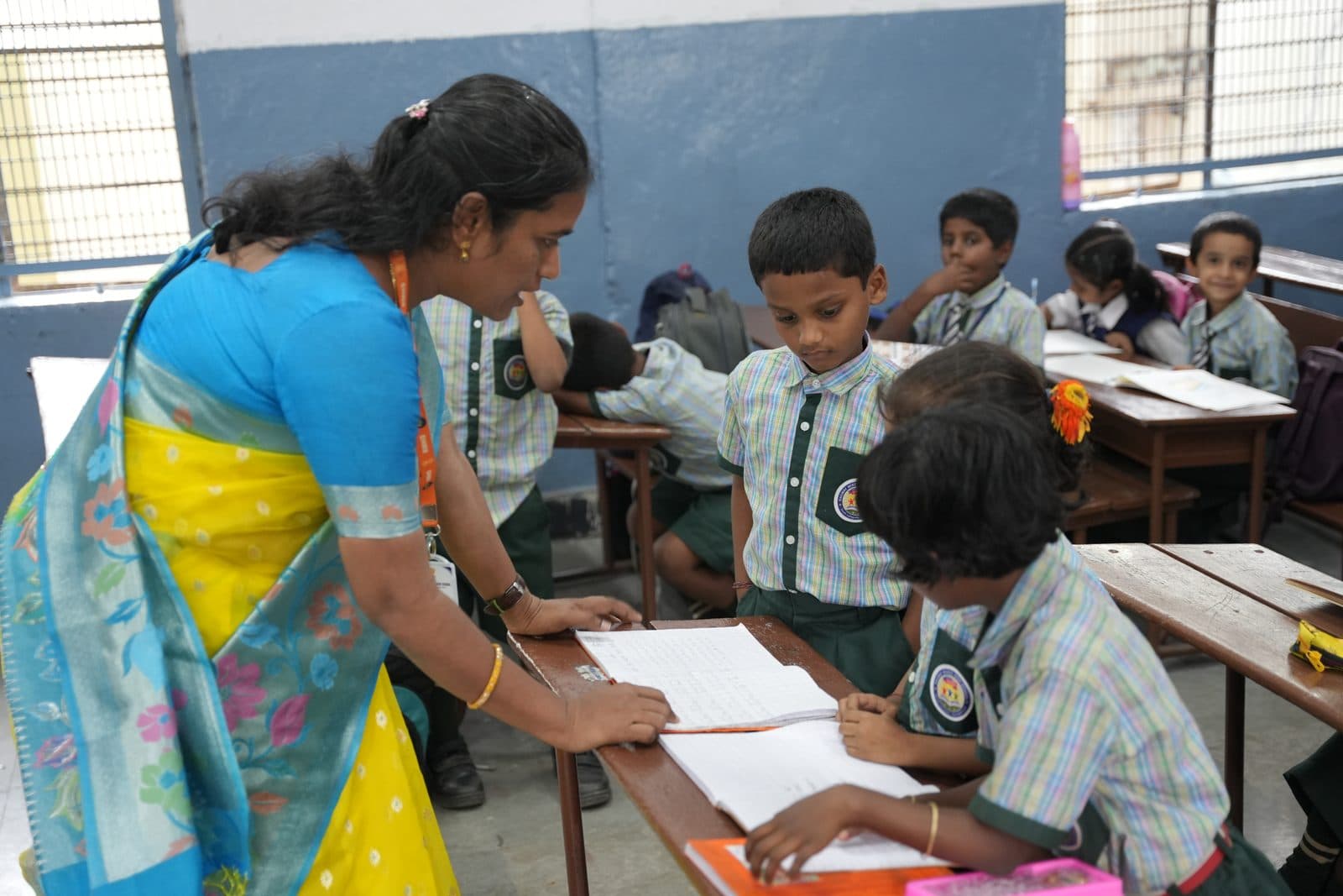A teacher leaning over a desk to guide two young students through their written work