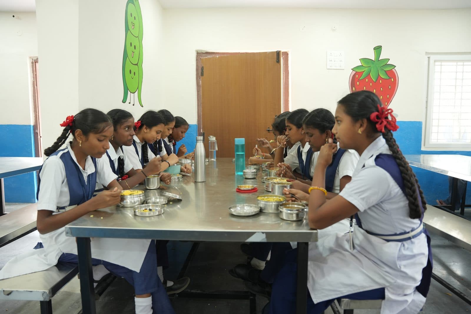Students seated along the dining table eating from stainless steel tiffin boxes