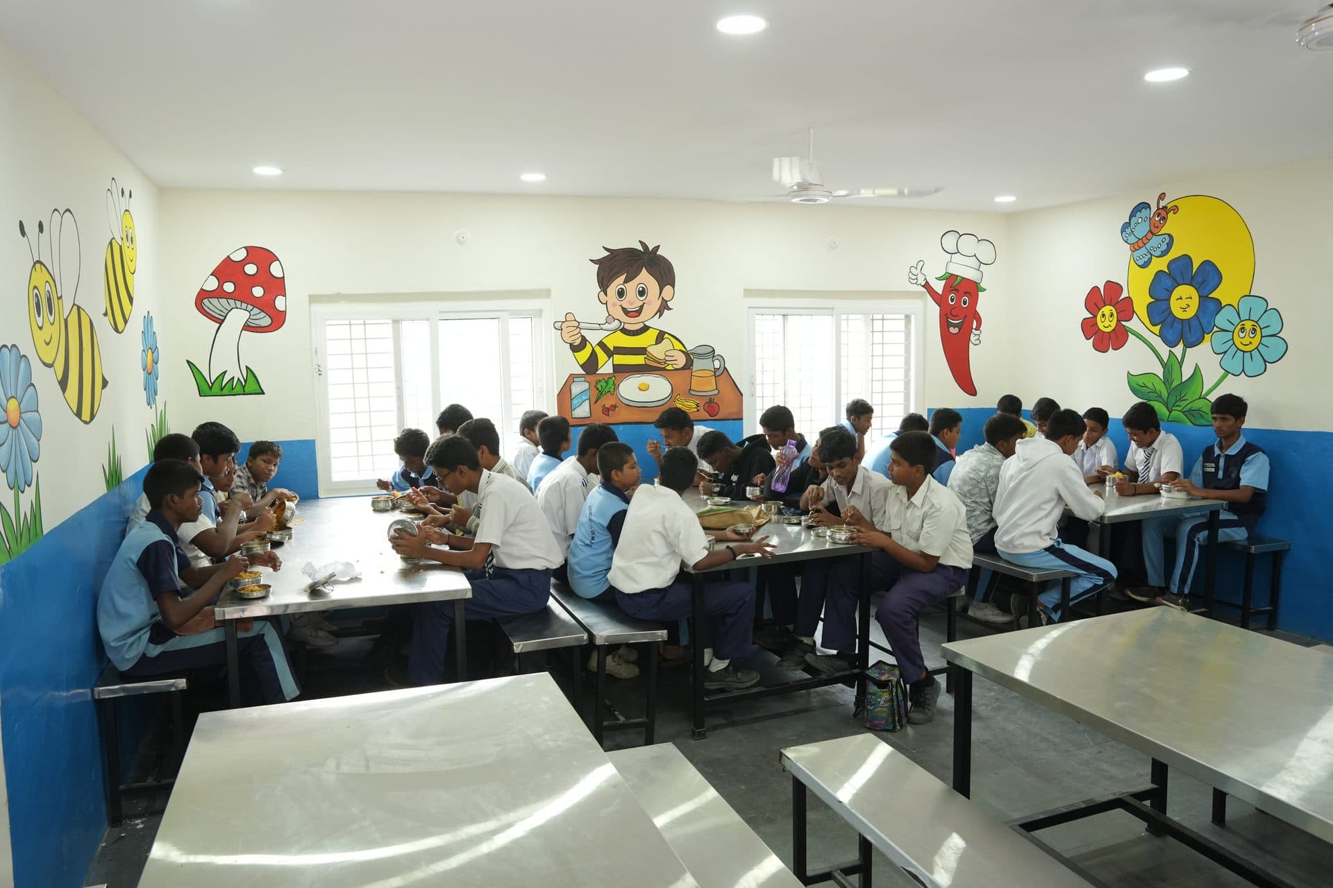 The Tripti dining hall filled with students eating lunch at long steel tables, with painted food murals on the walls