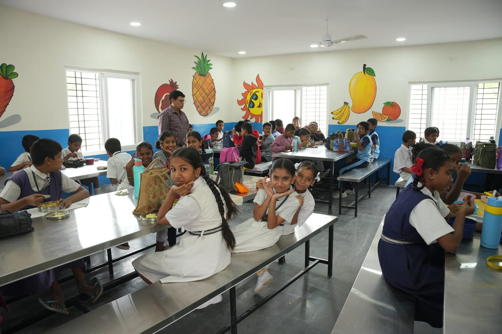 Students eating lunch together at a steel dining table while a teacher supervises